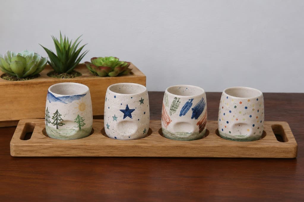 A set of four hand-painted ceramic thumb tumblers on a wooden flight board, each tumbler painted differently by a child